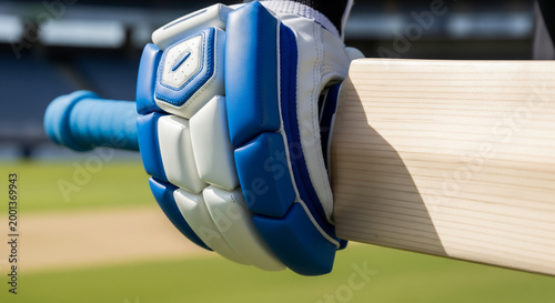Cricket glove holding bat on green field in stadium, Close-up view of blue and white sports equipment