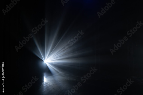 Bright light beam projecting from a dark background, creating dramatic rays and shadows on a smooth floor surface in a dimly lit environment