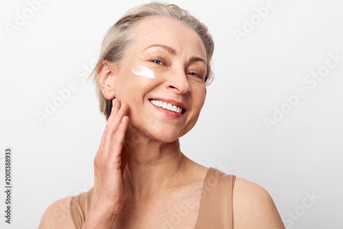 Smiling mature woman with short grey hair applies white face cream on cheek. Natural skincare concept with healthy glowing skin and anti aging beauty routine on white studio background.