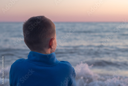 A little boy in a warm blue sweatshirt  sitting on the shore and looking at the sea on a cool spring day