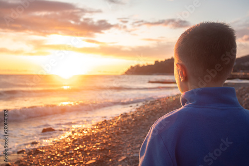 A boy in a blue sweatshirt standing on the seashore and admiring the sunset.