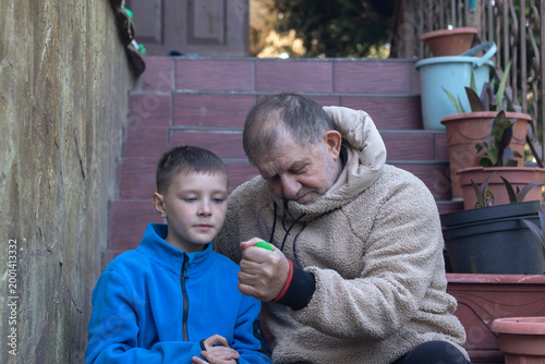 An elderly man showing his grandson  how he working out with a wrist expander while sitting on the steps in front of his house.