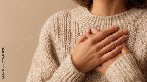 A woman's hands on her chest holding her heart to symbolize emotion, love, or vulnerability, wearing a cozy beige knitted sweater, warm and intimate setting