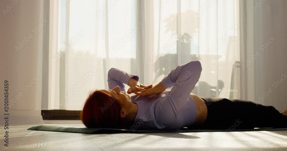 Fototapeta premium In a bright living room, a woman engages in stretching and pilates exercises on a mat. The sunny space promotes a healthy morning routine filled with movement.