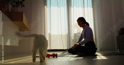 Cuadro en lienzo A woman sits on a fitness mat in her living room, wearing headphones as she engages her cat with a toy, creating an enjoyable and active environment indoors