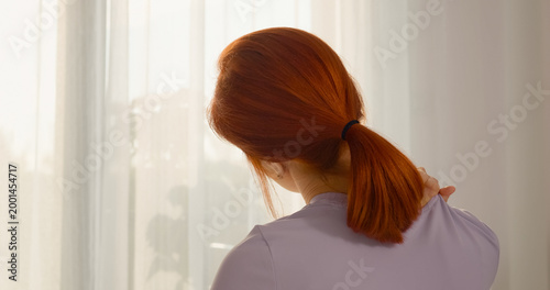 Photos In a cozy living room, a redhead woman is gently massaging her neck to alleviate muscle pain