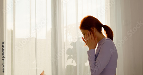 Obraz na płótnie A redheaded woman experiencing headache pain tries to alleviate her discomfort in the living room of her home, dealing with symptoms of migraine and neck strain