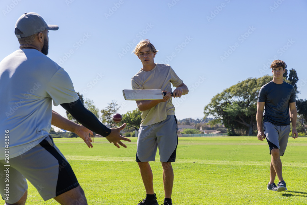 Obraz premium Three men playing cricket in sportswear on field, batter swinging flat wooden bat, ball airborne