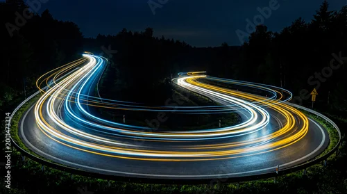 Obraz  z motywem Dynamic long exposure photograph of vibrant car light trails on a curving road at night through a dark forest.
