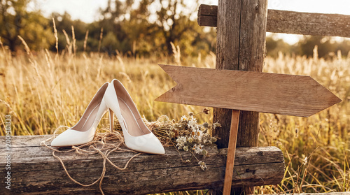 Obraz na płótnie White bridal high heels and a small dried bouquet on a rustic wooden fence post in a sunny field with an arrow sign