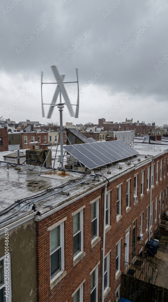 Fototapeta premium Vertical axis wind turbine and solar panels mounted on flat roof of urban brick rowhouse