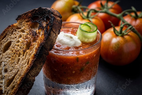 Spanish gazpacho cold tomato soup served in glass with toasted bread and cucumber garnish on dark background