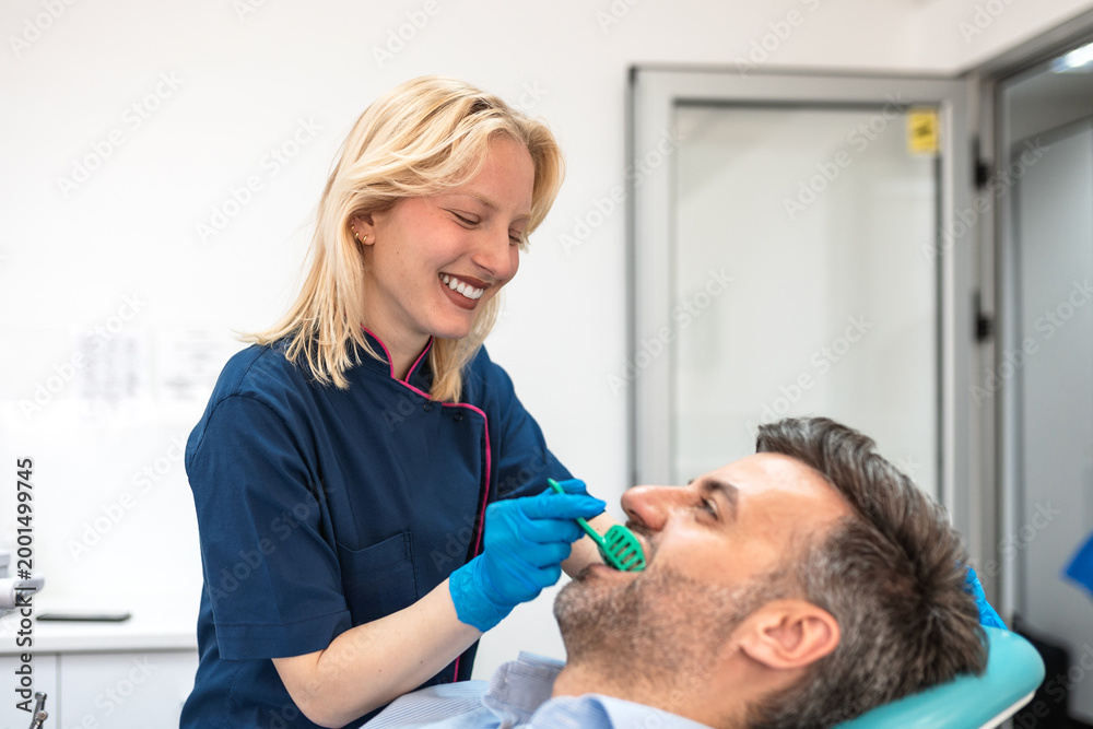 Fototapeta premium Smiling female dentist taking dental impression of male patient in clinic. Healthcare and medicine concept. Young woman doctor using tray for tooth mold, orthodontics or dental implant procedure.