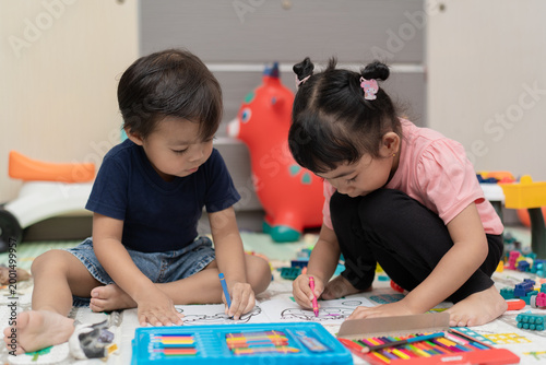 Two young Asian children coloring together in a book on a floor mat.