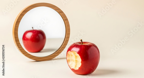A bitten red apple sits on a smooth surface, reflected in a round wooden-framed mirror