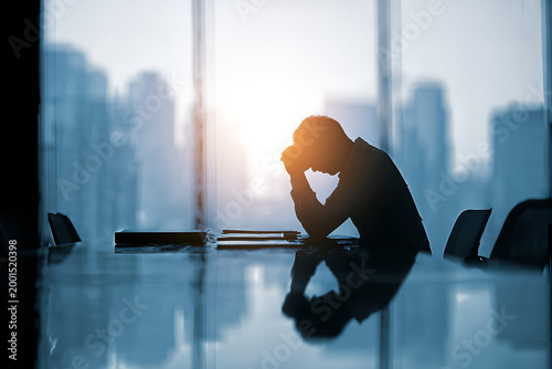 Silhouetted business executive with head in hands in a boardroom at sunrise — corporate stress, burnout and workplace anxiety against a city skyline