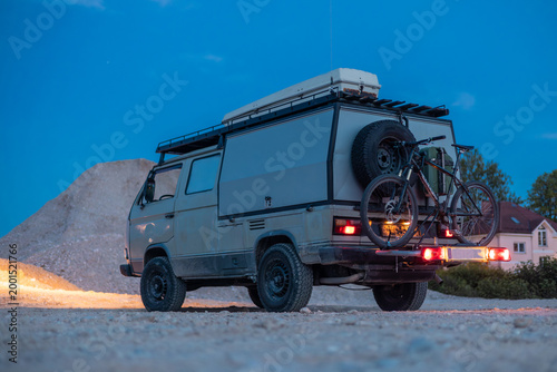 A rugged off-road camper van parked on gravel at dusk, equipped with a roof rack, mountain bike, and spare tire. A modern green-accented building stands in the background under a blue twilight sky.