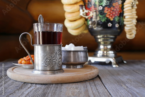 Fotografie On a wooden table on the background of log wall there is a glass of black tea in a metal cup holder next to a samovar with bagels