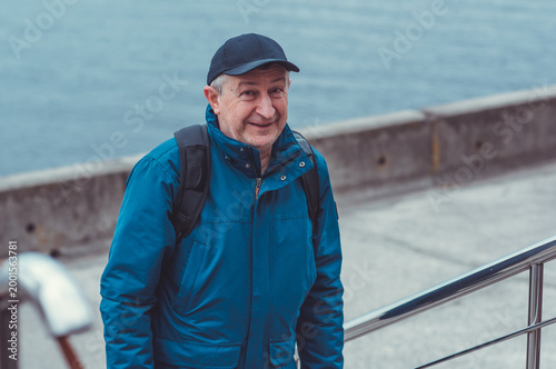 Smiling Senior Man in Blue Jacket and Cap by Riverside. Approachability and Well-being Concept