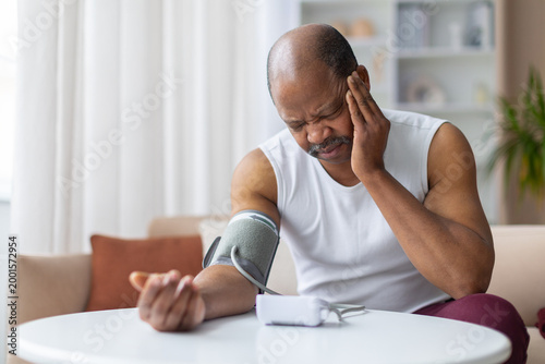 Distressed mature African American man wearing blood pressure cuff while holding head in pain. High blood pressure, hypertension, or headache medical health concept at home