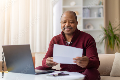 Картината върху платно Pleased mature African American man reading paper document beside laptop at home, smiling contentedly