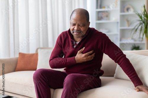 Bild auf Leinwand Mature African American man clutching his chest in pain while sitting on sofa at home