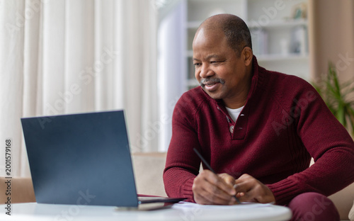 Smiling mature African American man taking notes while working on laptop at home desk. Remote work, studying, or financial planning lifestyle concept in living room interior