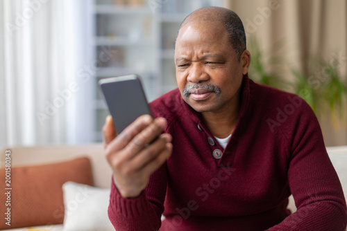 Vászonkép Confused mature African American man squinting and frowning at smartphone screen at home