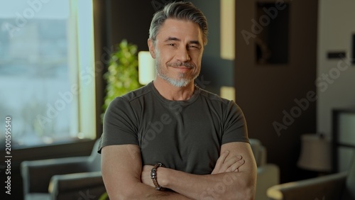 Portrait of a relaxed, smiling mature adult male with arms crossed in a stylish interior. Happy confident fit muscular mid adult older male in 50s with friendly smile. .
