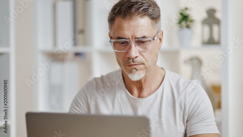 Middle aged casual man sitting at desk, looking at laptop, concentrating. Entrepreneur working from comfort of home office. Businessman listening on video meeting, making business decision, thinking.