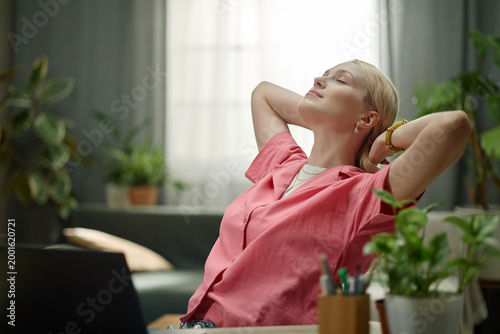 Photos Caucasian young adult woman leaning back and stretching at desk beside laptop, taking break during home office work