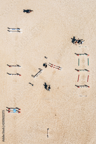 Aerial view of horses and riders training on a sandy show jumping arena with colorful obstacles