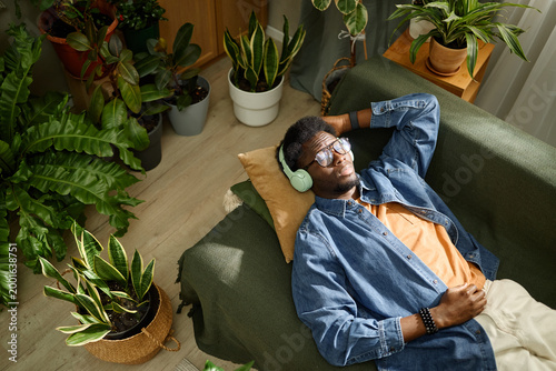 High angle shot of young adult Black man resting on sofa, listening through headphones among houseplants, representing mindfulness and home self care. Ideal for wellness campaigns