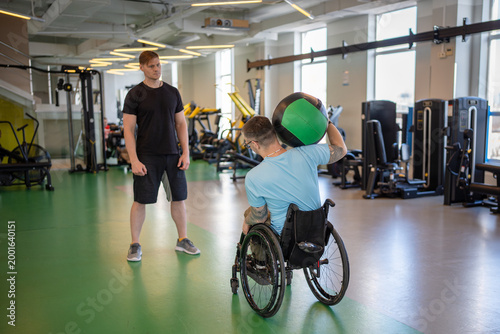 Photos Professional fitness trainer watching disabled sportsman in wheelchair on rehabilitation gym training, man doing upper body exercises using heavy medicine ball to build back and neck muscles strength