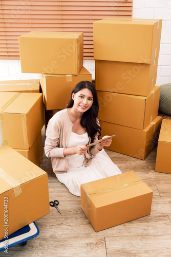 Smiling woman sitting on the floor among stacked cardboard boxes, packing orders with tape and scissors, preparing shipments from a home workspace