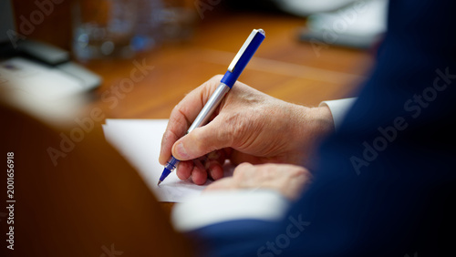 Elderly executive signing official paperwork in a boardroom. Close up of an older man hand writing a letter or endorsing a legal business deal on a wooden table. Photo