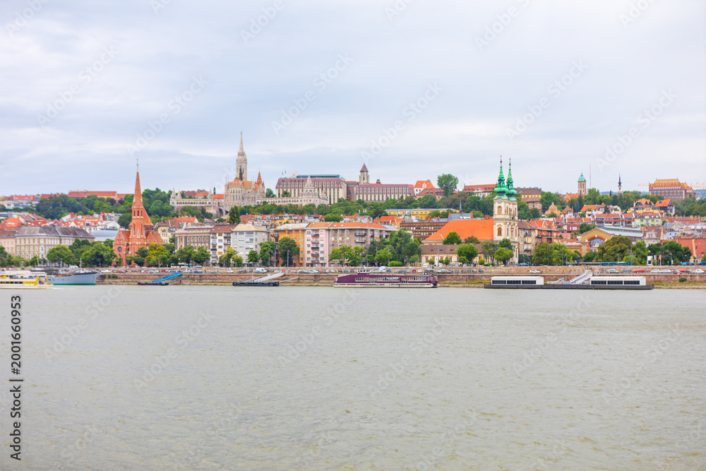 Obraz premium Budapest historic Buda side unfolds along the Danube River under overcast sky. Skyline features the spires of Matthias Church and Fisherman's Bastion rising above colorful riverside buildings