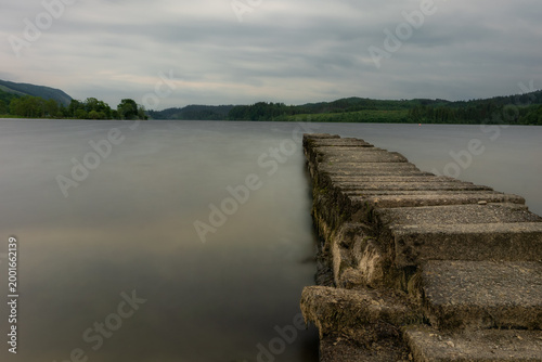 Long exposure of a tranquil loch at Kinlochard, near Loch Lomond, Scotland, showcasing a weathered concrete pier leading into still waters under an overcast sky.