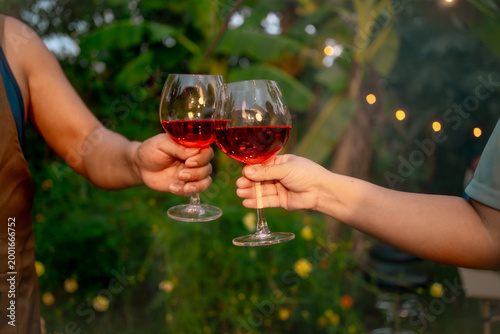 Close-up of two people clinking glasses of red wine outdoors in a garden at dusk with warm bokeh lights, celebrating together.
