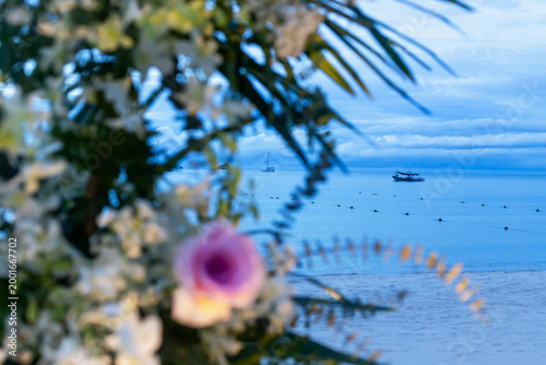 Romantic sunset. Serene tropical beach scene featuring floral decorations in foreground, with boats visible on calm water in distance, under cloudy sky during sunset