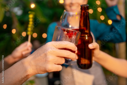 Friends clinking beer bottle, wine glass, and cocktail at outdoor party with warm bokeh string lights, shallow depth of field, celebrating together.