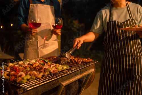 Two people in aprons grilling kebabs and steak on outdoor barbecue at night, holding red wine glasses, warm lighting, casual dinner party atmosphere.