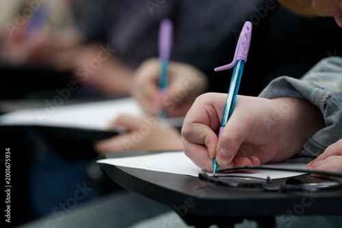 Group of students taking a test at their desks. Selective focus on a hand holding a pen over a blank sheet. Ideal for university lectures, homework, and diligence themes. Photo