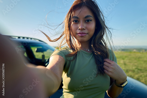 Young asian woman smiling while taking a candid selfie outdoors, capturing a moment of freedom and happy travel during a road trip with her car under a clear blue sky