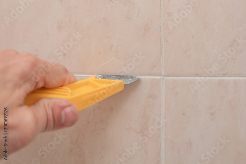Male hand cleaning seams from the old sealant between ceramic tiles joints. Tile joint preparation before grouting using handheld tool