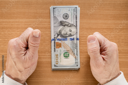 Hands securing stacked hundred dollar bills on wooden table, male hands flanking banded bundle, warm natural light highlighting texture of skin and paper, closeup of cash