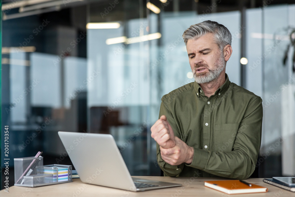 Obraz premium Senior man working on a laptop at an office desk, gripping his aching wrist due to discomfort, showing symptoms of carpal tunnel syndrome or workplace injury from overuse