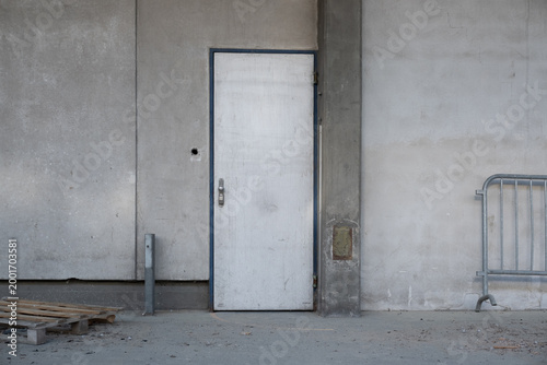 Minimalist white metal door on a gray concrete industrial wall with a pallet and metal fence