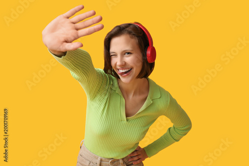 Beautiful young woman in modern headphones showing tongue on yellow background