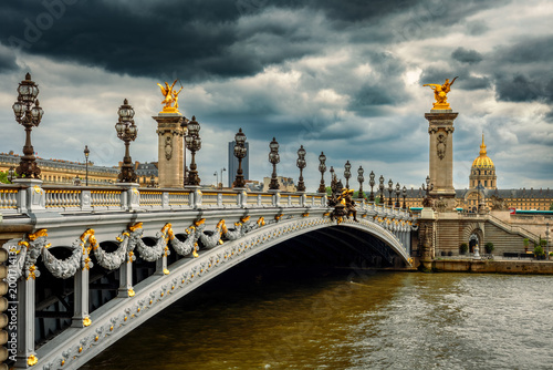 Pont Alexandre III bridge in Paris, with gilded sculptures and lampposts, under a dramatic and stormy sky over the Seine River.